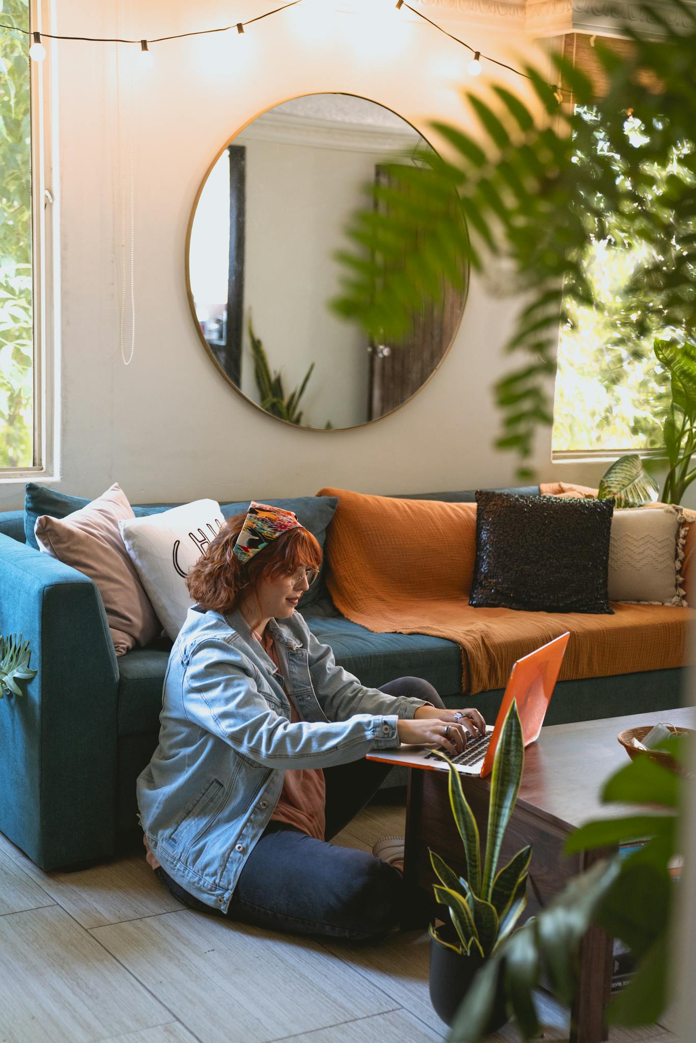 Woman with red hair working from home on a laptop in a stylish living room surrounded by plants.